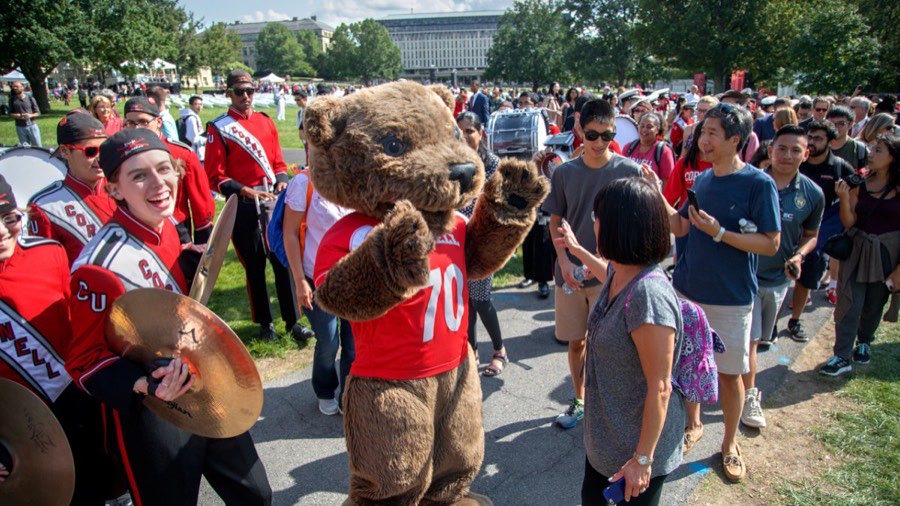 Marching band members with cymbals walking beside the Cornell bear mascot through a crowd on the quad