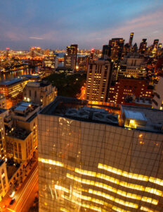 A city skyline at dusk, with lit office windows reflected in a glass tower