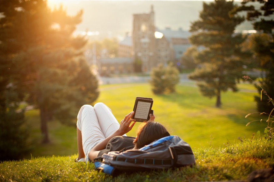 A student lying on a grassy hillside reading a tablet, with the campus clock tower behind